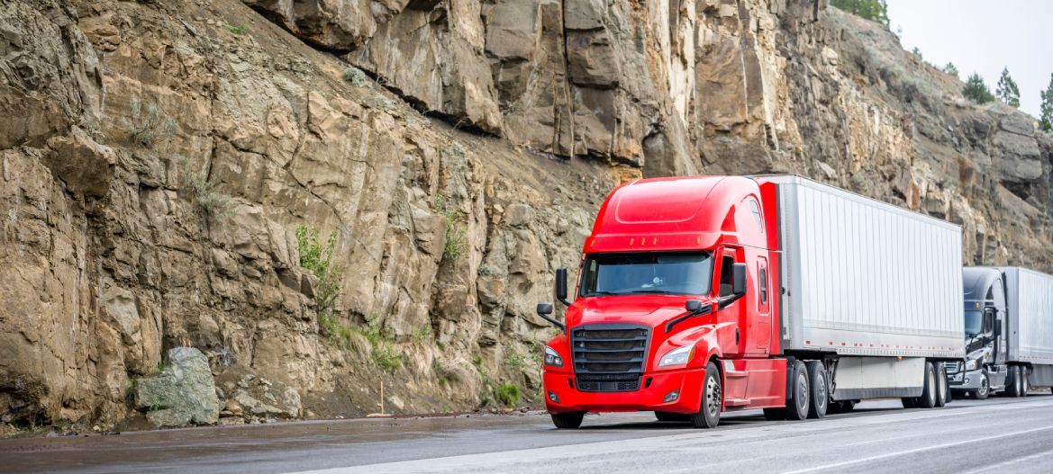 Group of big rigs semi trucks tractors transporting cargo in different semi trailers standing off road in a line near a stone cliff take a break at the pass on top of a mountain range in California