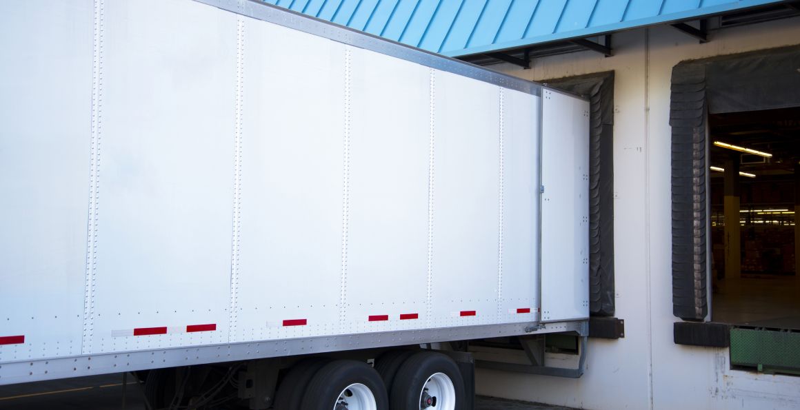 White long semi trailer with an open door stand close in the warehouse dock with a green sloping roof to unload a cargo delivered to this location.