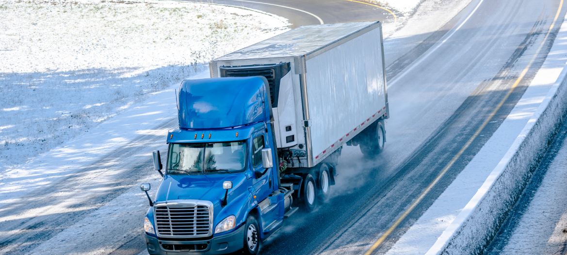 Big rig blue bonnet day cab semi truck with roof spoiler and refrigerator unit on the front wall of reefer semi trailer transporting commercial cargo driving on the turned winter wet road with snow