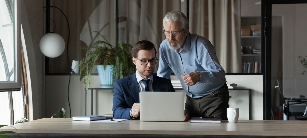 Senior businessman wearing glasses mentor instructing training new employee, using laptop, helping with corporate software, two colleagues working on online project together, discussing strategy