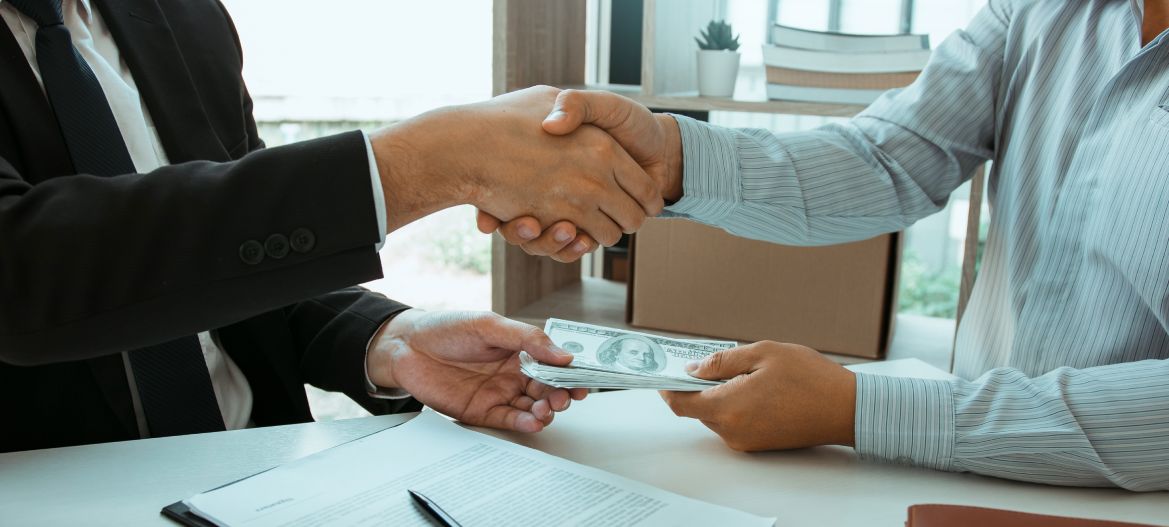 Two corporate businessmen shaking hands while one man places money on document in office room with corruption concept.